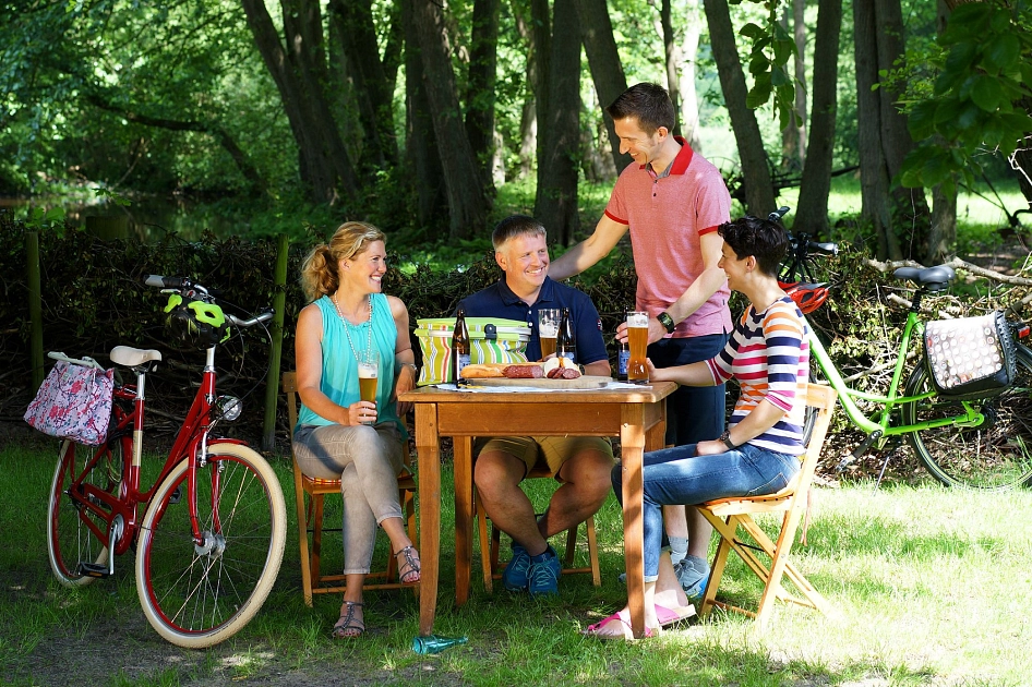 Vier Radfahrer machen im Wald eine Pause und Essen und Trinken dort an einem TIsch.