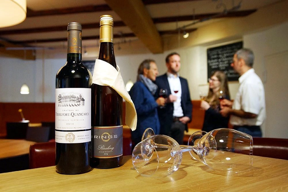 Andreas_Herrmann_Weinprobe_Höltingmühle Zwei Flaschen Wein und Weingläser im Vordergrund, im Hintergrund steht eine Gruppe von Menschen und trinkt Wein. © Andreas Herrmann