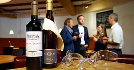 Zwei Flaschen Wein und Weingläser im Vordergrund, im Hintergrund steht eine Gruppe von Menschen und trinkt Wein.